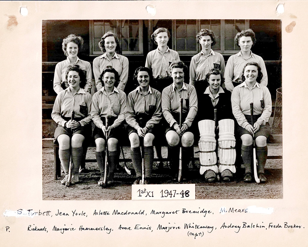 Black and white photograph of a smiling women's hockey team posed in two rows, the back row standing and the front row seated holding hockey sticks.