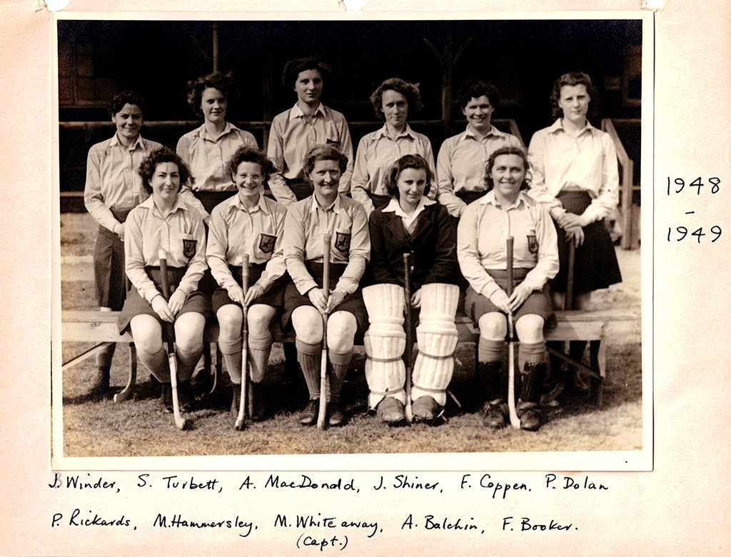 Sepia toned black and white photograph of a smiling women's hockey team posed in two rows, the back row standing and the front row seated holding hockey sticks.