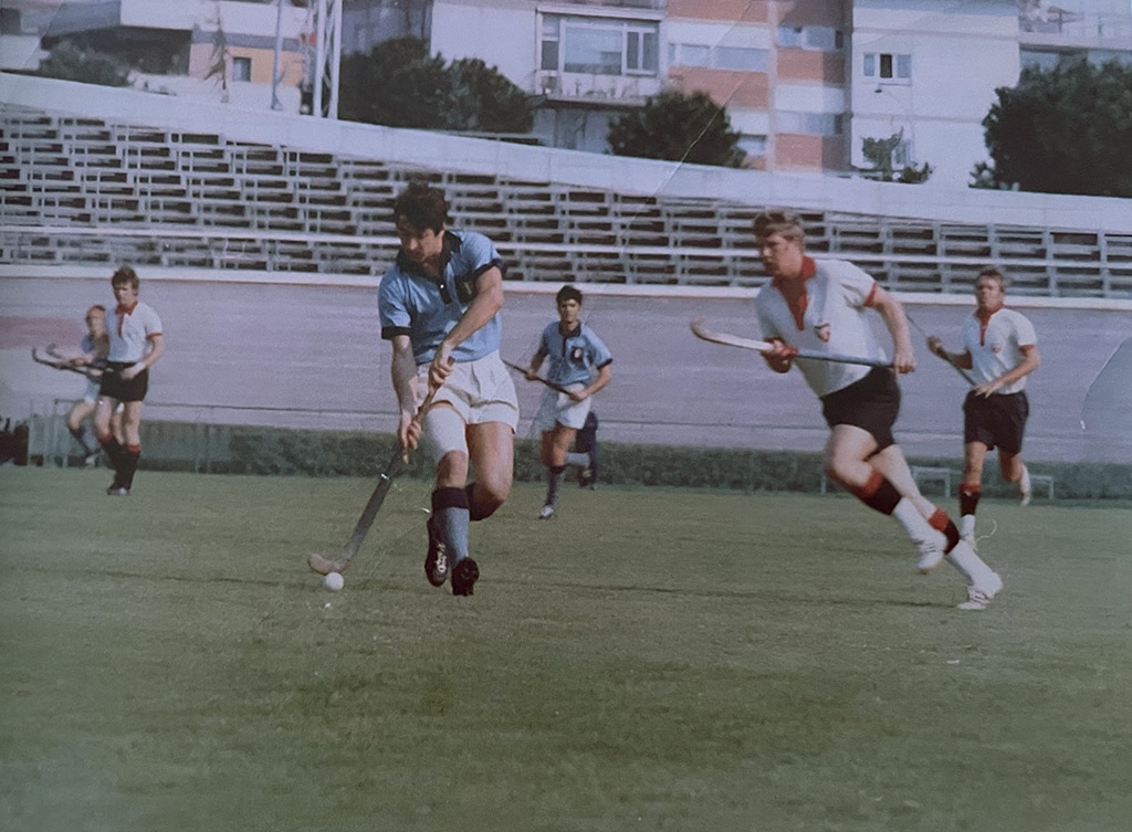 Action photograph of a young male hockey player wearing a light blue shirt and white shorts dribbling a hockey ball at pace. Other players wearing white chase him.