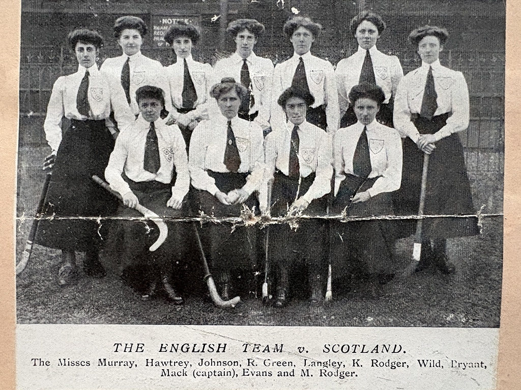 A team of women posed in two rows holding hockey sticks. They are wearing long-sleeves blouses with long ties and long skirts.