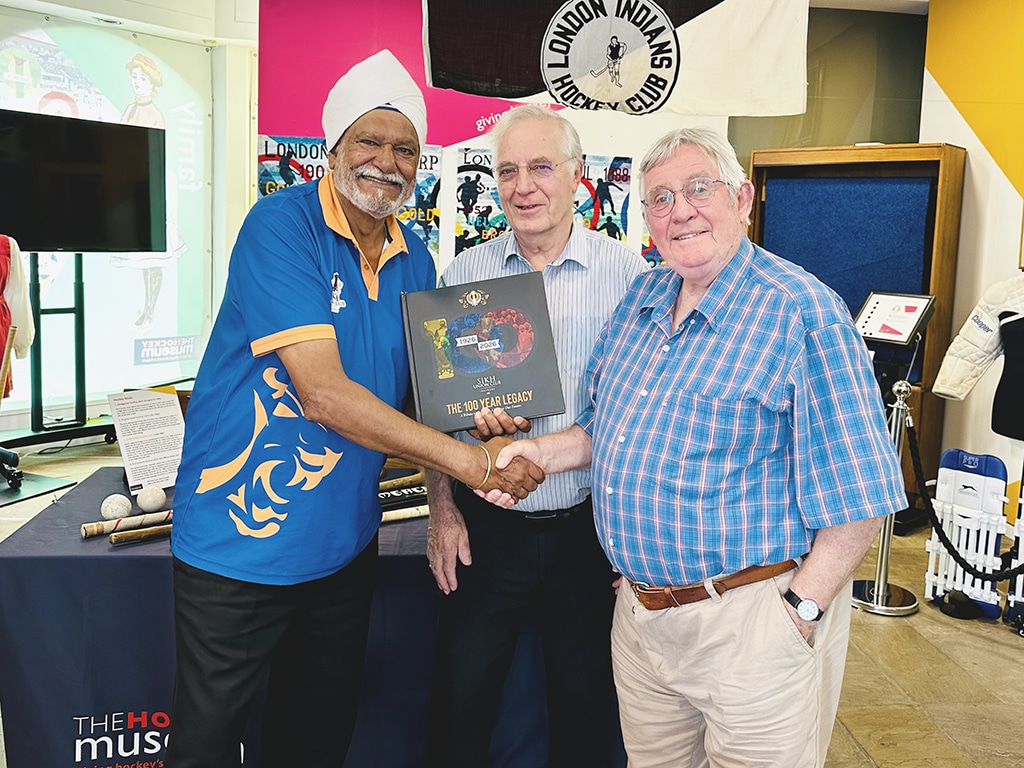 Three elderly men hold a large black book between them while shaking hands.