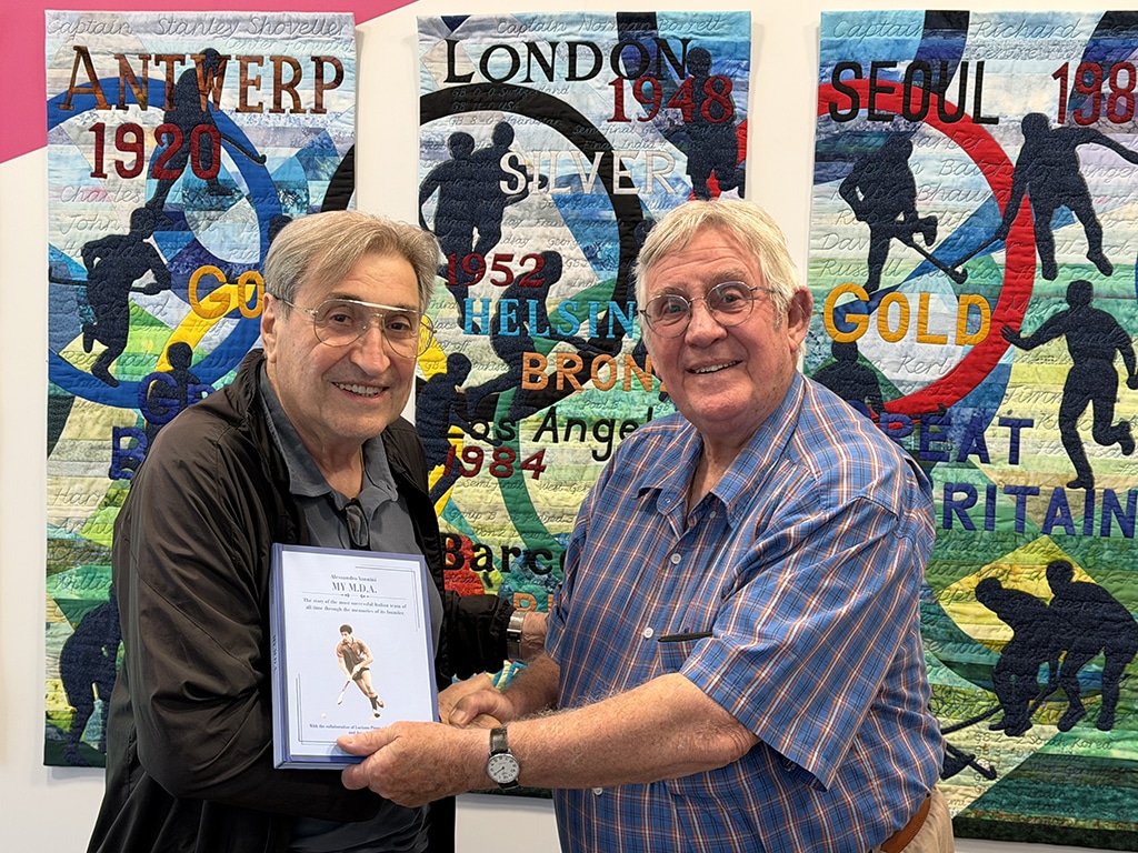 Two elderly men wearing spectacles shake hands whilst passing a blue covered book between them.