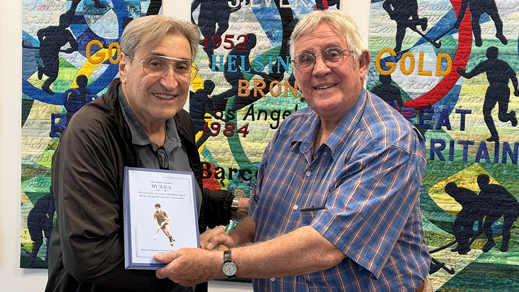Two elderly men wearing spectacles shake hands whilst passing a blue covered book between them.