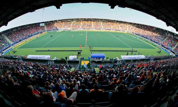 Elevated panoramic view of a hockey stadium with a green pitch and spectators filling the stands around the field.