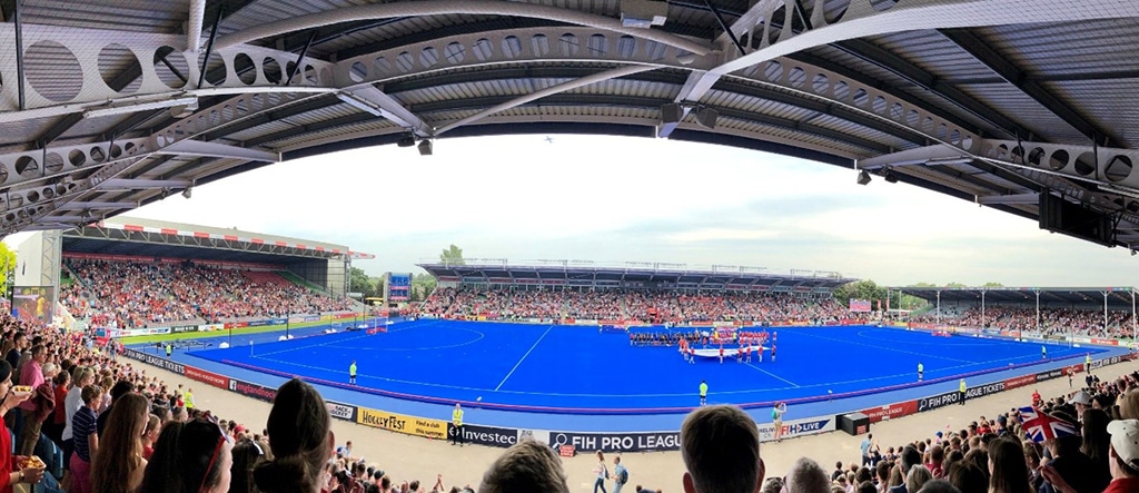 Panoramic view of a hockey match venue with a blue pitch, packed stands, and teams gathered near the centre of the field for the national anthems.