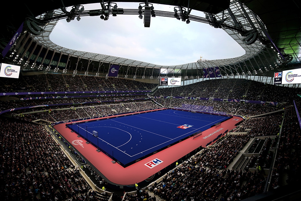 A blue hockey pitch inside a vast stadium with thousands of spectators filling the surrounding stands.