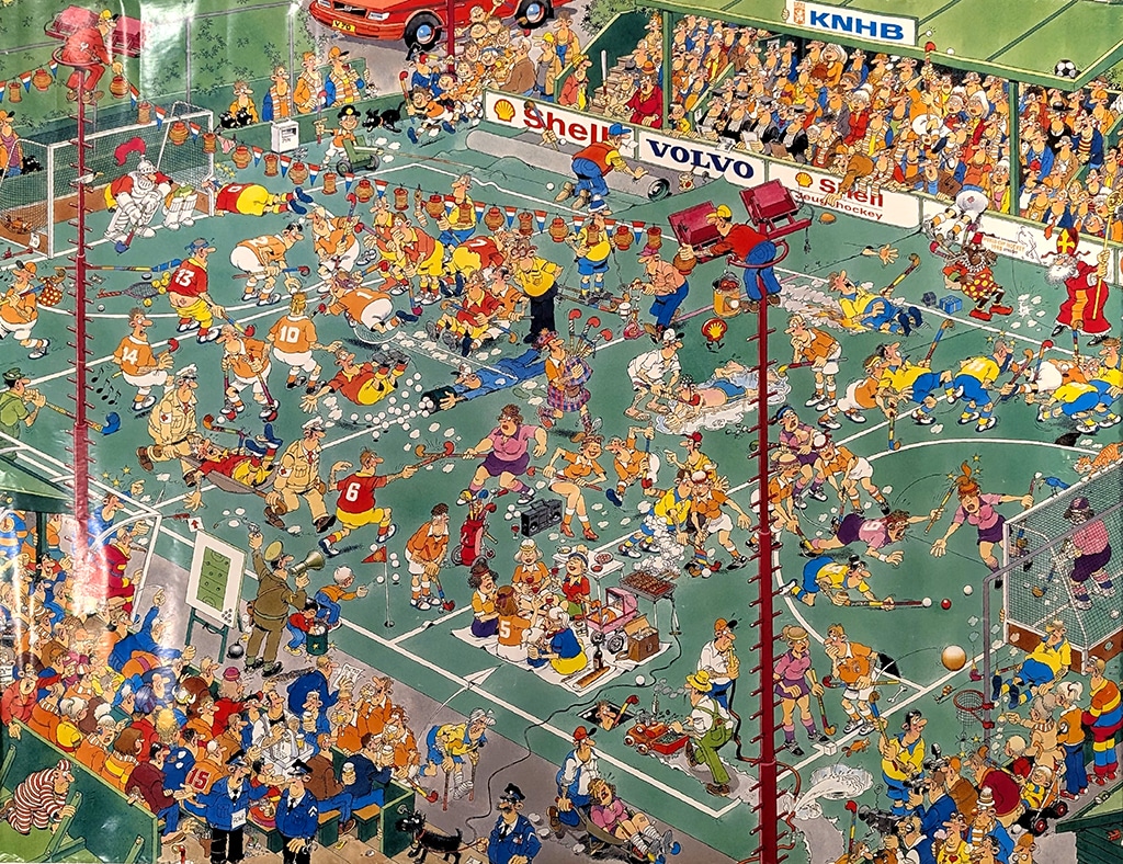 Large, colourful cartoon-style poster depicting a hockey stadium. The hockey pitch is crowded with dozens of players, officials, media crews, ground staff, mascots and spectators engaged in a wide variety of humorous and chaotic scenes. Players in orange, yellow, blue and red uniforms are shown running, tackling, falling, shooting and colliding across multiple areas of the field. Hockey balls, sticks and equipment are scattered throughout the scene. Around the pitch, television cameras, photographers, groundskeepers, vendors and maintenance vehicles add to the chaotic activity.