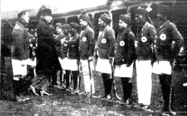 Black-and-white photograph of a hockey team standing in a line with hockey sticks while being greeted by officials on the side of a grass hockey pitch.