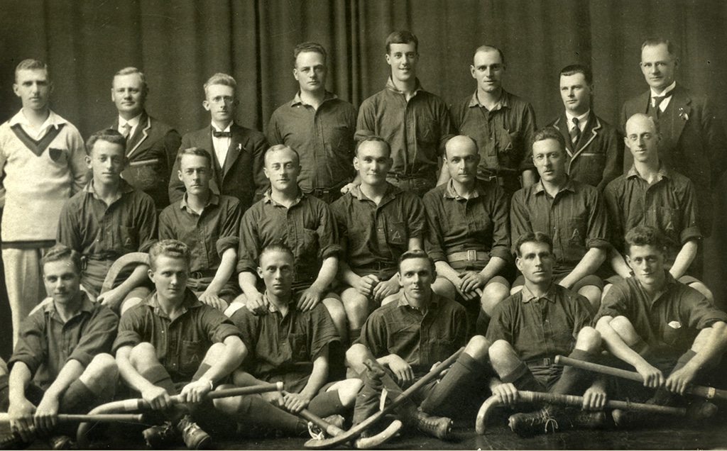 Black and white photograph of a squad of men arranged in three rows in front of a fabric backdrop. The front row and sitting cross-legged holding hockey sticks.