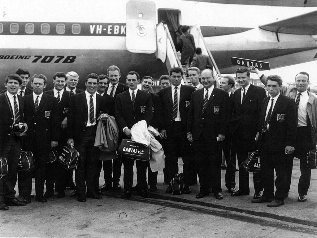 Black-and-white group photograph of a hockey touring team standing on an airport tarmac in front of a Qantas Boeing 707B aircraft. Team members wear matching Great Britain blazers and hold travel bags.