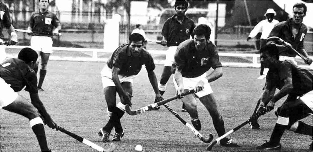 Players compete for possession of the ball during a hockey match on a grass pitch, with several players clustered around the ball. The black-and-white photograph captures fast-paced action at close range.