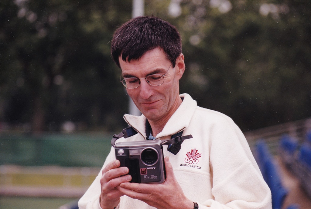 Person wearing glasses and a light-coloured fleece examines a compact Sony digital camera while standing outdoors. Trees and sports venue features are blurred in the background.