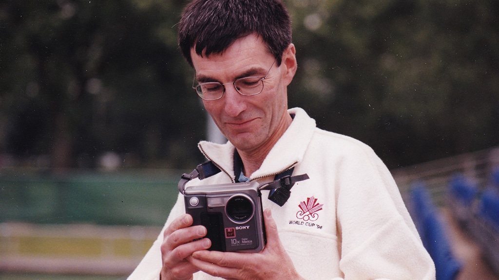 Person wearing glasses and a light-coloured fleece examines a compact Sony digital camera while standing outdoors. Trees and sports venue features are blurred in the background.