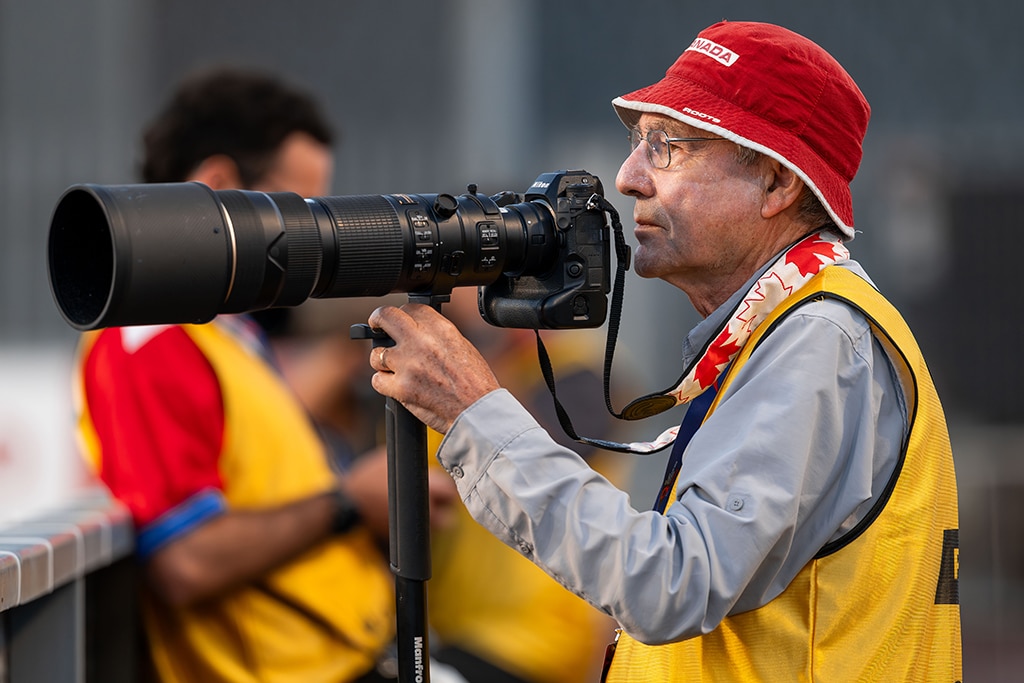 An accredited sports photographer wearing a red hat and a yellow media bib uses a professional camera fitted with a large telephoto lens at the side of a sports venue.