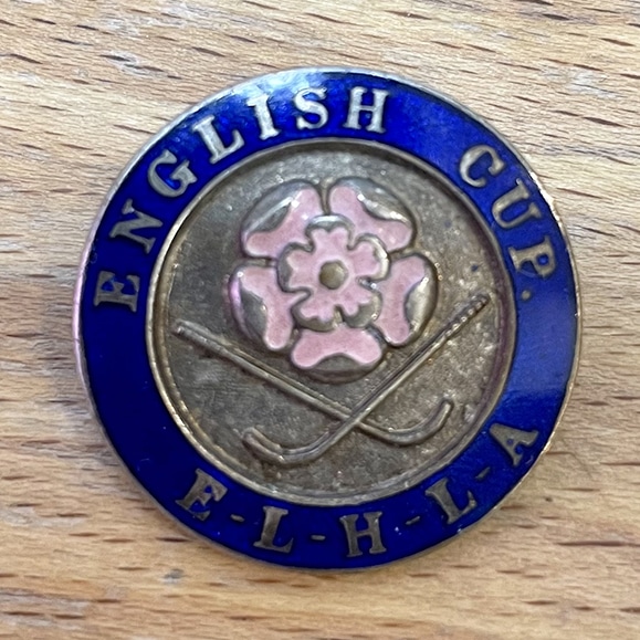 A circular pin badge with a central design of crossed hockey sticks below a Lancashire rose, ringed by a royal blue band with the words, "English Cup, ELHLA".