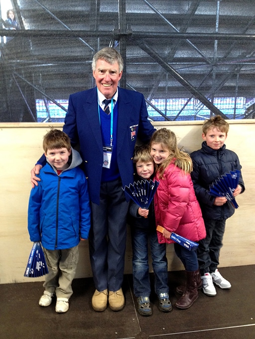An older adult man wearing a blue blazer and accreditation lanyard stands with four children in an indoor venue. The children hold blue folding fans and wear colourful jackets.
