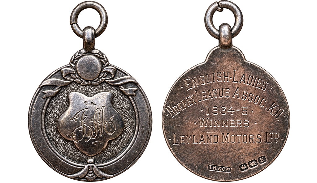 The front and reverse of a circular metal medal. The design on the front includes a central shield with the initials JM, above which is a ribbon and wreath motif. The reverse is inscribed, "English Ladies Hockey Leagues Association Knock Out, 1934-35. Winners Leyland Motors Ltd."