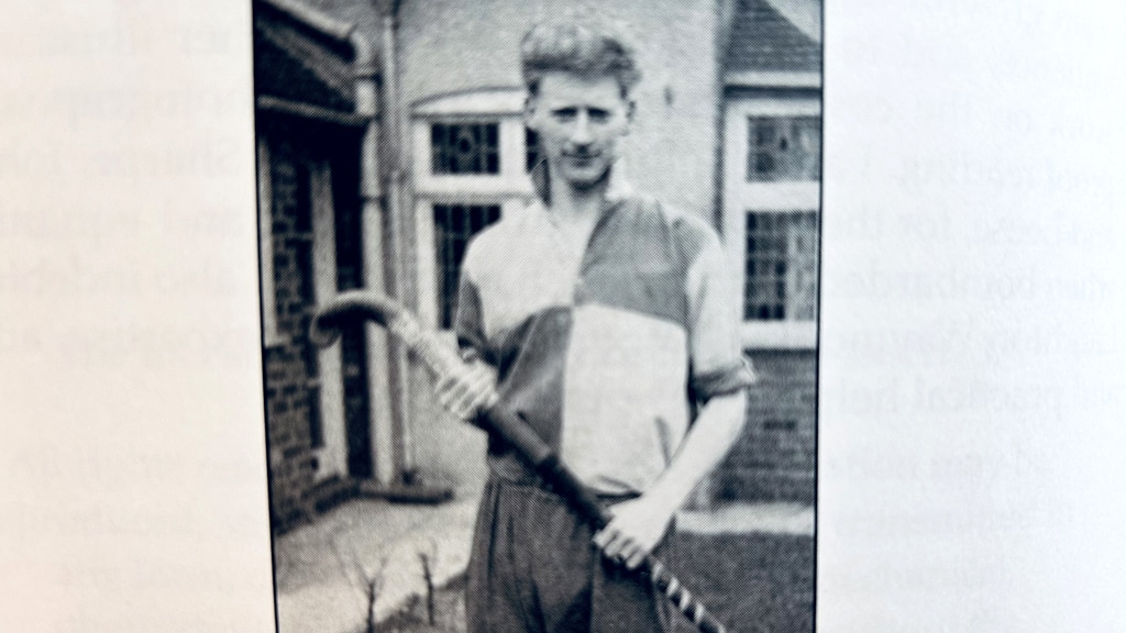 Black-and-white photograph of a hockey player standing outdoors and holding a field hockey stick diagonally across the body. The player is wearing a short-sleeved quartered sports shirt and matching shorts. Behind the player are brick buildings with windows and a paved area leading to a doorway.