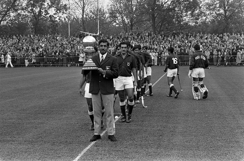 Black-and-white photograph of a field hockey pitch. A player wearing a blazer walks along the centre line carrying the Hockey World Cup trophy, followed by several Pakistan players in national team uniforms. A large crowd lines the far side of the field, with trees and goalposts visible in the background.