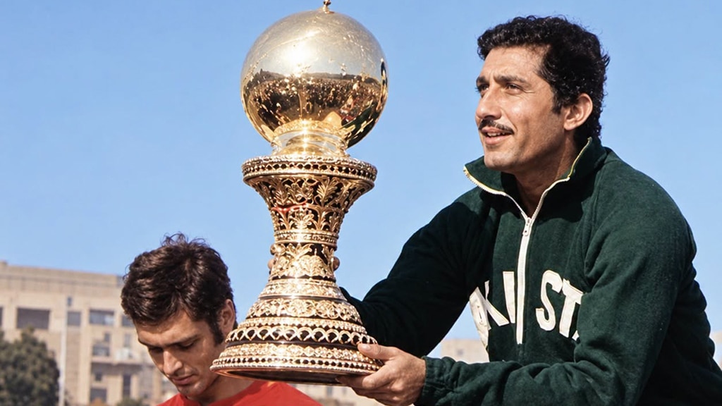 Colour photograph of a field hockey trophy presentation. A Pakistan player in a green tracksuit top and white shorts holds an ornate gold World Cup trophy with a large globe-shaped top.