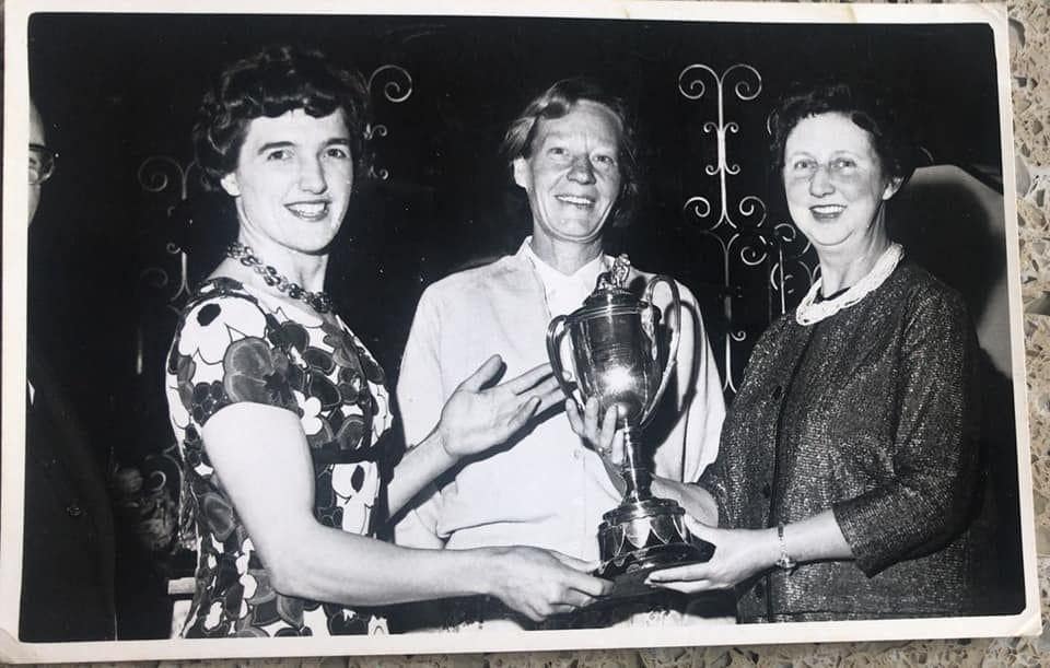 Black-and-white photograph showing three women at an indoor trophy presentation. A large two-handled cup trophy is being held between the individuals.
