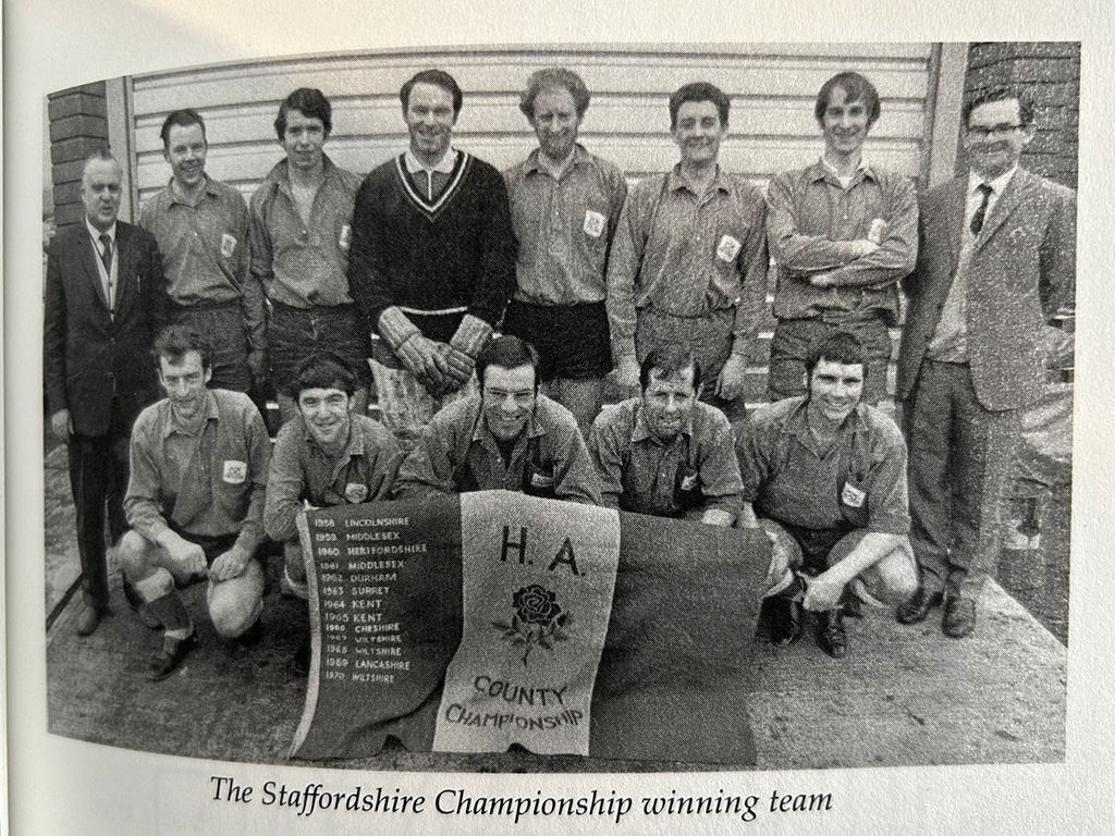 Black-and-white team photograph showing eleven male hockey players arranged in two rows. Six are standing behind and four are crouching at the front, with two additional people standing at either end of the group in suits. A large banner in the foreground reads “H.A. County Championship” and includes a rose emblem and a list of previous county winners.