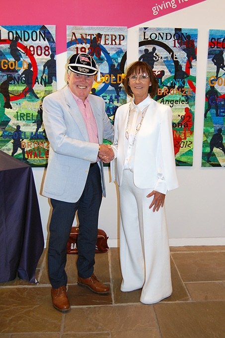 A man and a woman stand shaking hands at an indoor event, posed in front of a colourful quilted artwork featuring references to Olympic Games and medal achievements. The man wears a light-coloured jacket and commemorative cap, while the woman wears a white suit. A display table is visible to the side and stone flooring is visible in the foreground.