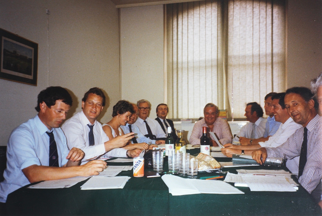 A group of middle-aged men in shirts and ties and one woman sit around a large table. There are wine, soft drinks and sandwiches on the table.