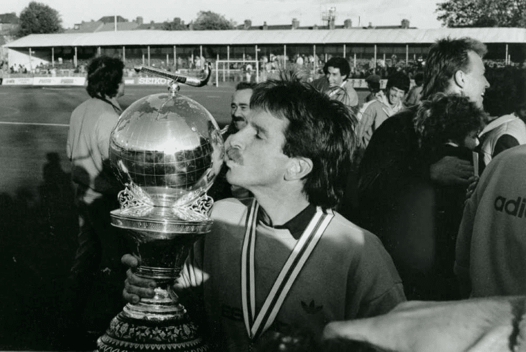 A man wearing a medal holds and kisses the Hockey World Cup trophy. Other players, officials and spectators stand nearby, with the stadium grandstand visible in the background.