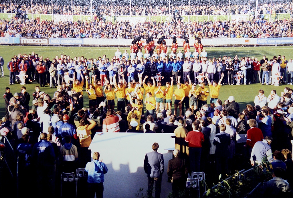 A team of players wearing yellow shirts and green shorts stand together on the pitch with their arms raised as they celebrate winning the 1986 Hockey World Cup. Surrounded by photographers, officials and opposition teams, the winning team is presented to a packed stadium crowd.