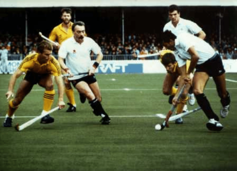 Players from two international hockey teams in white and yellow kits contest possession of the ball during a match. Several players crouch low with sticks extended as the ball sits near the touchline, with a crowded grandstand visible in the background.