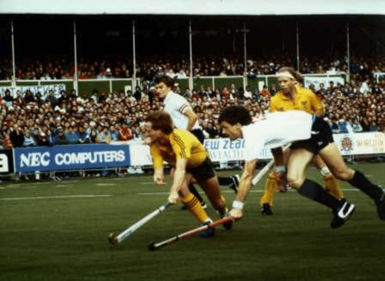 A player in a yellow kit drives the ball forward along the sideline while a player in a white kit stretches to challenge. Other players follow the play, with spectator stands packed with fans behind advertising boards around the pitch.