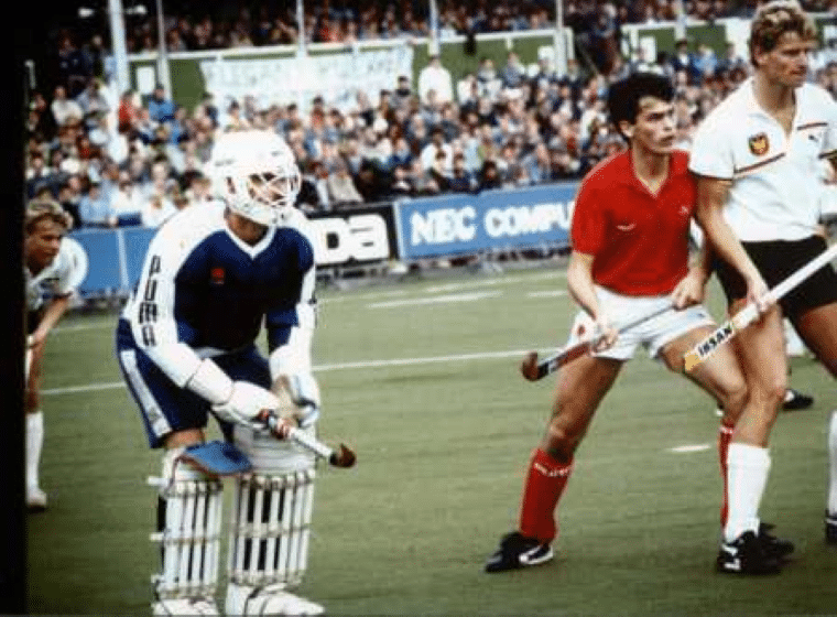 Players from opposing teams contest play inside the shooting circle during an international hockey match. A fully protected goalkeeper in blue and white kit, pads and helmet stands ready in front of goal while players in red and white kits watch the developing attack. A packed grandstand fills the background.