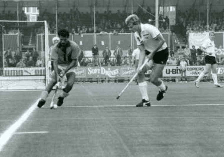 Two hockey players sprint along the pitch baseline as one controls the ball close to the line and the other pursues with stick extended. A goal and additional players are visible in the background, with spectators watching from the grandstand.