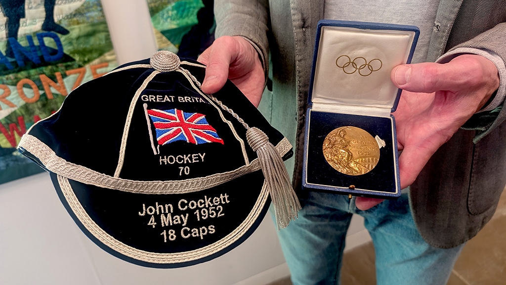 Close up of a navy blue cap with silver trim and tassel with the Great Britain flag stitched onto it, and a circular medal in a blue presentation box with Olympic rings on the inside fabric.