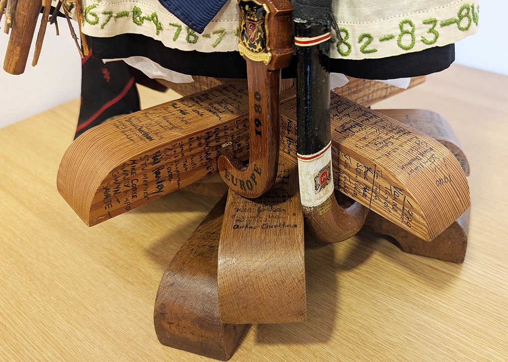 Close-up of the base of a commemorative mascot: several curved wooden slats are arranged in a star shape. The sticks are covered with handwritten names and dates, with two miniature hockey sticks positioned vertically at the centre.