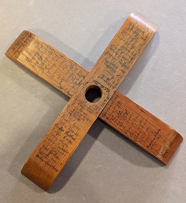 Cross-shaped stand made from overlapping wooden slats, viewed from above. The varnished wood is densely covered with handwritten signatures, and dates surrounding a central mounting hole.