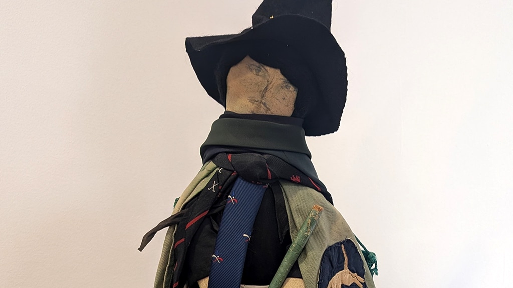 Close-up of the face of a handcrafted hockey-themed witch doll, wearing a tall black pointed hat and layered fabric garments decorated with badges and hockey club ties.