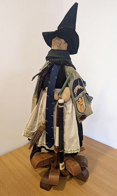 Handcrafted hockey-themed witch doll, wearing a tall black pointed hat and layered fabric garments decorated with badges, ties and embroidered text. The figure stands on a wooden base which is covered with handwritten signatures and inscriptions.