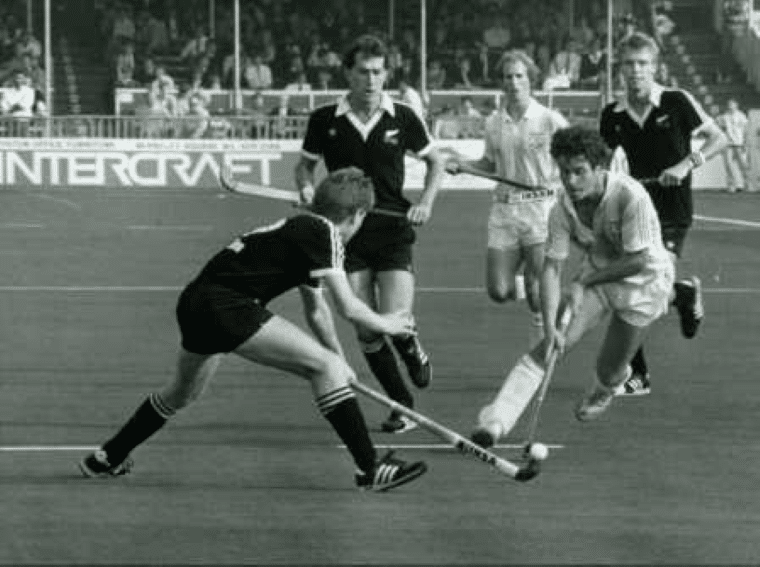 A player in a light blue kit controls the ball while moving forward, challenged by a player in black uniform crouching low with a stick extended. Three other players follow the play, with spectators filling the stands in the background.