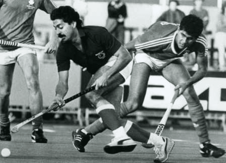 Two players lean forward and compete for the ball at close quarters, their sticks crossing near the ground. Another player is partially visible nearby, with advertising boards and spectators in the background.
