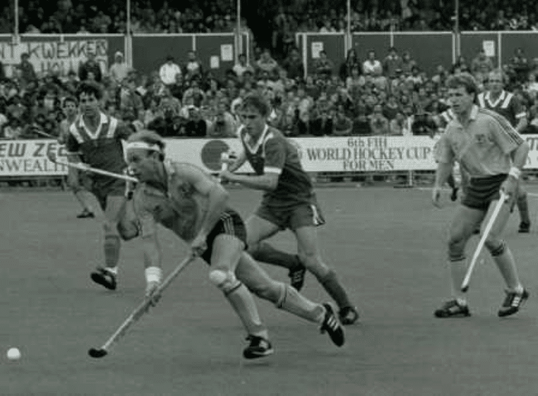 A player wearing a yellow shirt, green shorts and a headband runs forward with the ball while holding a hockey stick low to the ground. Three opposition players close in from behind, with spectators and World Cup signage visible around the pitch.