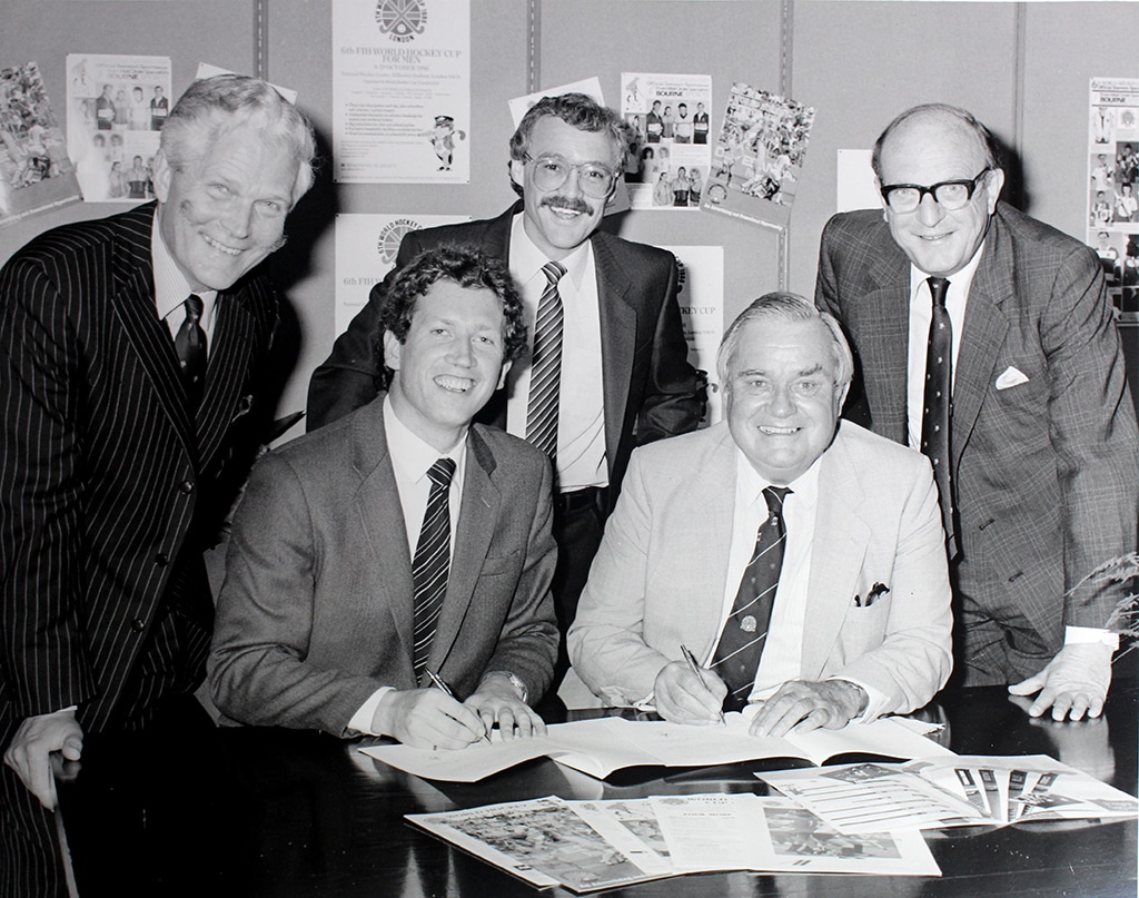 Five people in business suits gather around a table during a formal signing event. Two are seated and signing documents while three stand behind them. Promotional posters and printed materials related to a hockey tournament are displayed on the wall and table.
