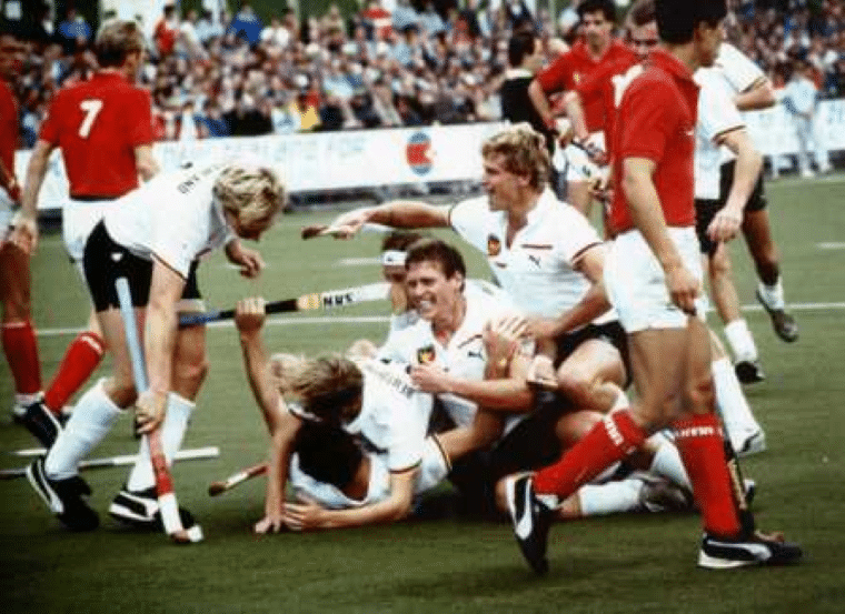 Players in white shirts and black shorts celebrate together on the pitch after scoring, with several teammates piled on top of one another. Opposing players in red shirts trudge away despondently nearby as spectators watch from the crowded stands.