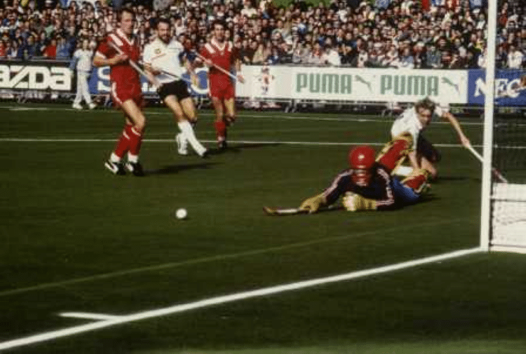 A fully protected goalkeeper wearing a red helmet and dark protective kit dives across the goalmouth as the ball rolls loose near the goal line. Several players in red and white uniforms run towards the play, with a packed crowd watching from the stands behind.